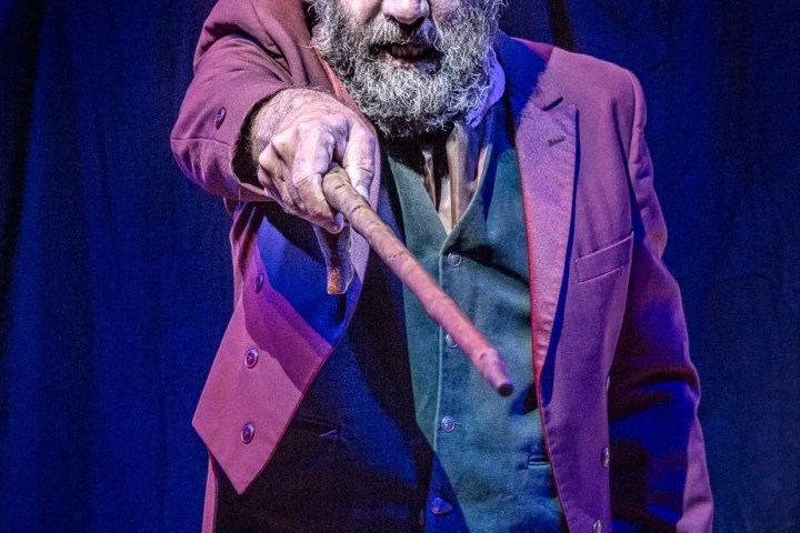 Man with a beard in a suit points a stick toward the camera on a dimly lit stage.