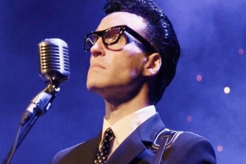 Man in suit with glasses singing into vintage microphone on stage.