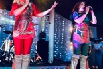 Two women in colorful outfits singing on stage with band instruments in the background.