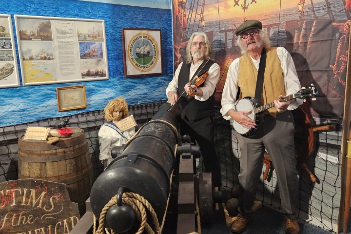 Two men playing guitar and banjo beside a cannon in a nautical-themed exhibit.