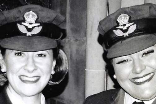 Two women smiling in vintage military uniforms with hats.
