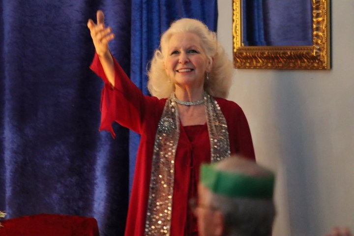 Smiling woman in red outfit gestures warmly, standing near blue curtain and gold-framed mirror.