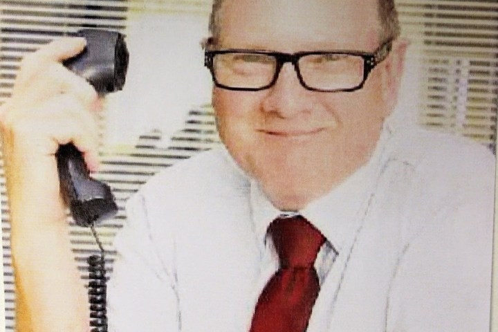 Man in glasses holding a telephone, smiling at a desk.