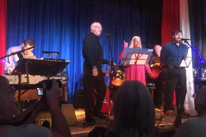 Band performing on stage with singers and keyboard player, audience watching.